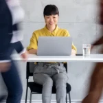 Happy Asian woman sitting at a desk with her laptop while coworkers rush around her.