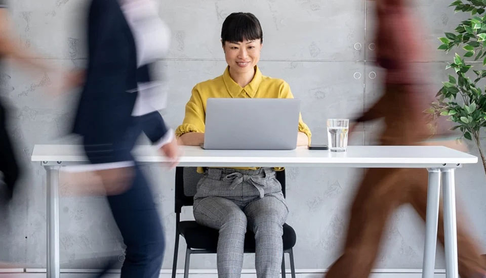 Happy Asian woman sitting at a desk with her laptop while coworkers rush around her.