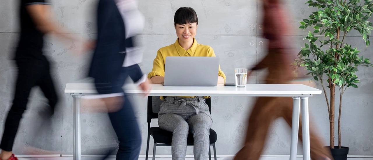 Happy Asian woman sitting at a desk with her laptop while coworkers rush around her.
